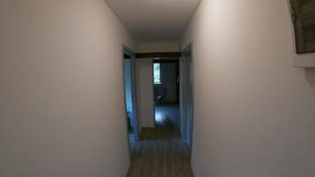 A narrow hallway with white walls and wooden flooring, leading to a room with a door.