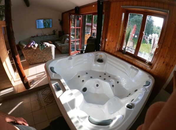 Un bain à remous dans une pièce en bois, équipée d’un téléviseur, d’un canapé et offrant une vue sur l’extérieur.