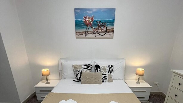 Una camera d'albergo con un letto, due lampade da comodino, un condizionatore d'aria a parete, un quadro di una bicicletta e un piccolo comodino con cassetti.