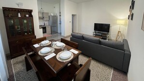 Zona pranzo con tavolo in legno apparecchiata per quattro persone, divano grigio, televisore e lampada.