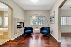 Ein modernes Wohnzimmer mit Holzdielenboden, zwei blaue Sessel, einem kleinen runden Tisch, einer Stehlampe und einem großen Fenster mit Jalousien.