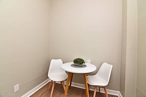 Ein Eckzimmer mit einem weißen runden Tisch und zwei weißen Stühlen, eine kleine Topfpflanze auf dem Tisch und eine weiße Wand mit sichtbarer Steckdose.