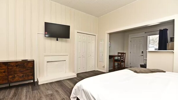 Une chambre à coucher avec un lit, un téléviseur fixé au mur, une commode en bois et une porte donnant sur une autre pièce.