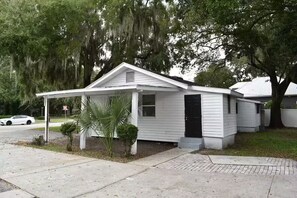 Ein kleines, eingeschossiges Haus mit einer Veranda, einer schwarzen Haustür und einem kleinen Vorgarten mit einer Palme und einem Busch.