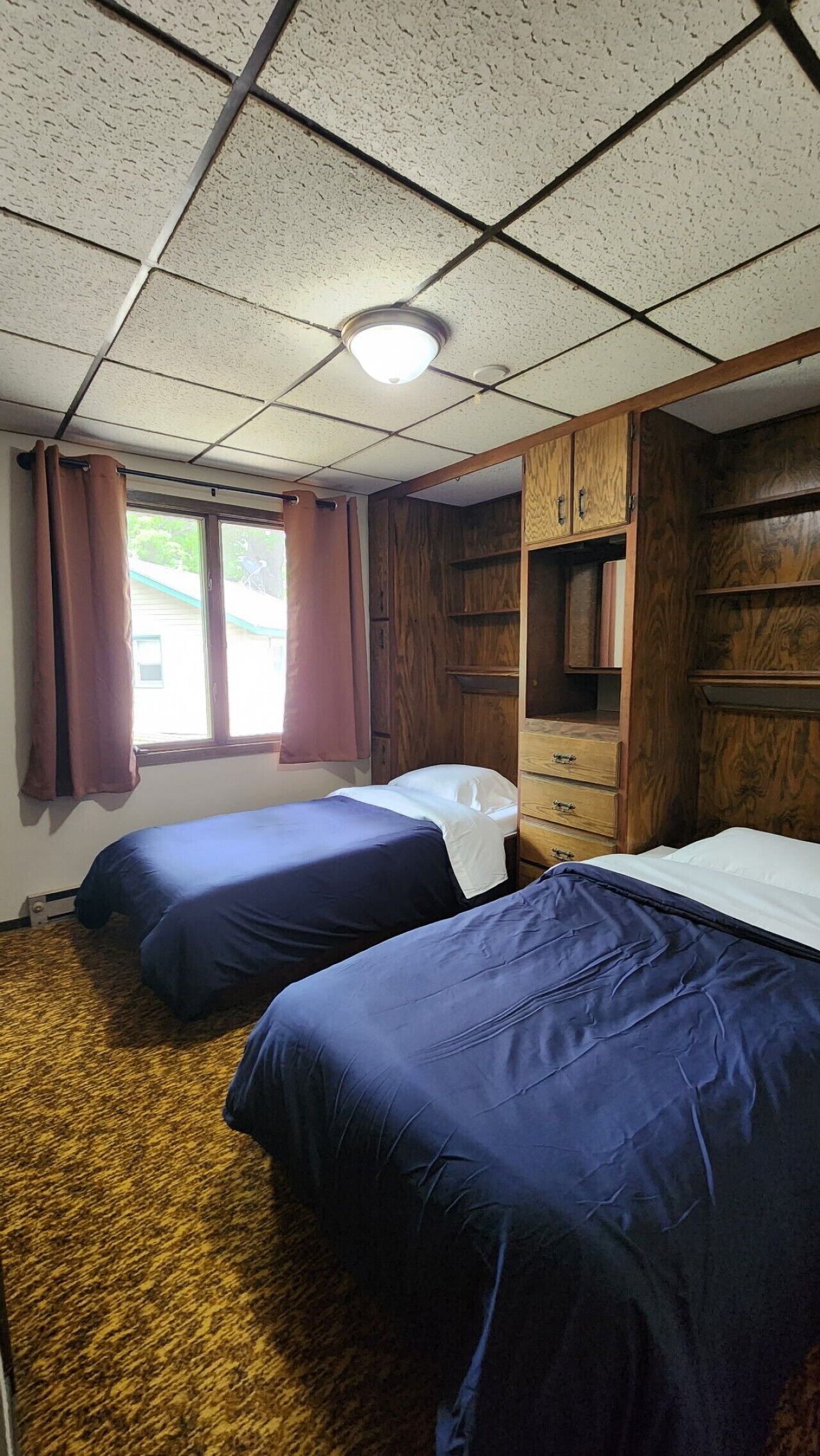 Una habitació amb dos llits, armaris de fusta, una finestra amb cortines i una làmpada al sostre.