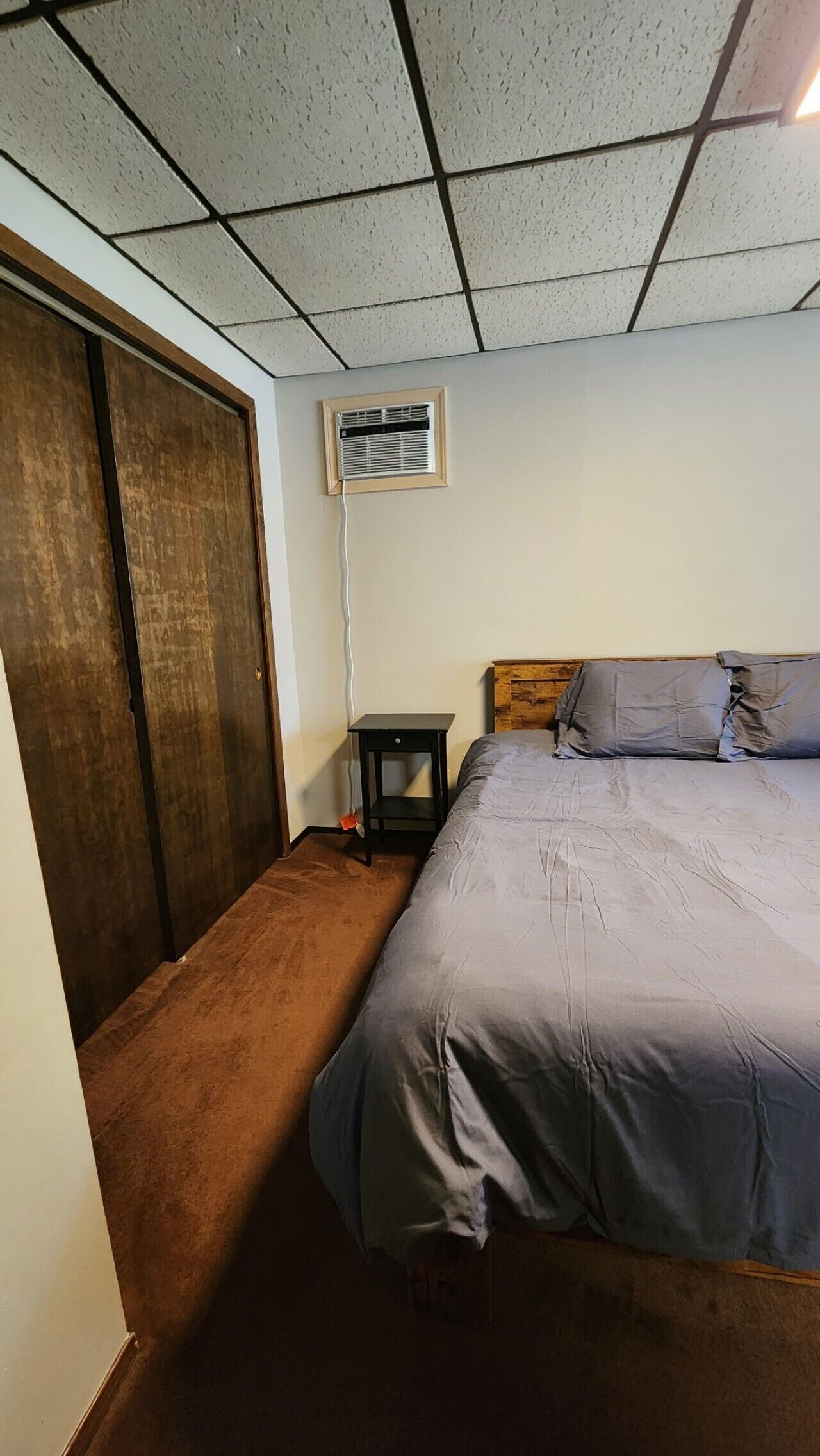 Un dormitorio con cama, cabecera de madera, mesita de noche, ventilación en la pared y una puerta.