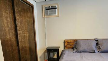 Un dormitorio con cama, cabecera de madera, mesita de noche, ventilación en la pared y una puerta.