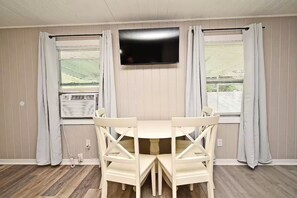 Ein Zimmer mit einem runden Tisch, vier Stühlen, einem flachbildfernseher an der Wand und zwei Fenstern mit Vorhängen.