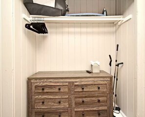 Eine hölzerne Kommode mit Schubladen, ein oberes Regal mit einer schwarzen Tasche und einem blauen Gegenstand, dazu eine Krücke, die an der Wand lehnt.