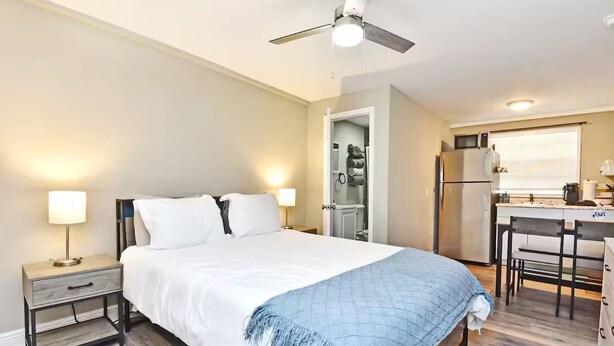 Een slaapkamer met een bed, nachtkastjes, een plafondventilator en een kitchenette met een koelkast en magnetron.
