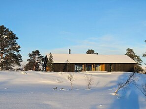 A wooden cabin with a snow-covered roof, surrounded by trees and a snowy landscape.