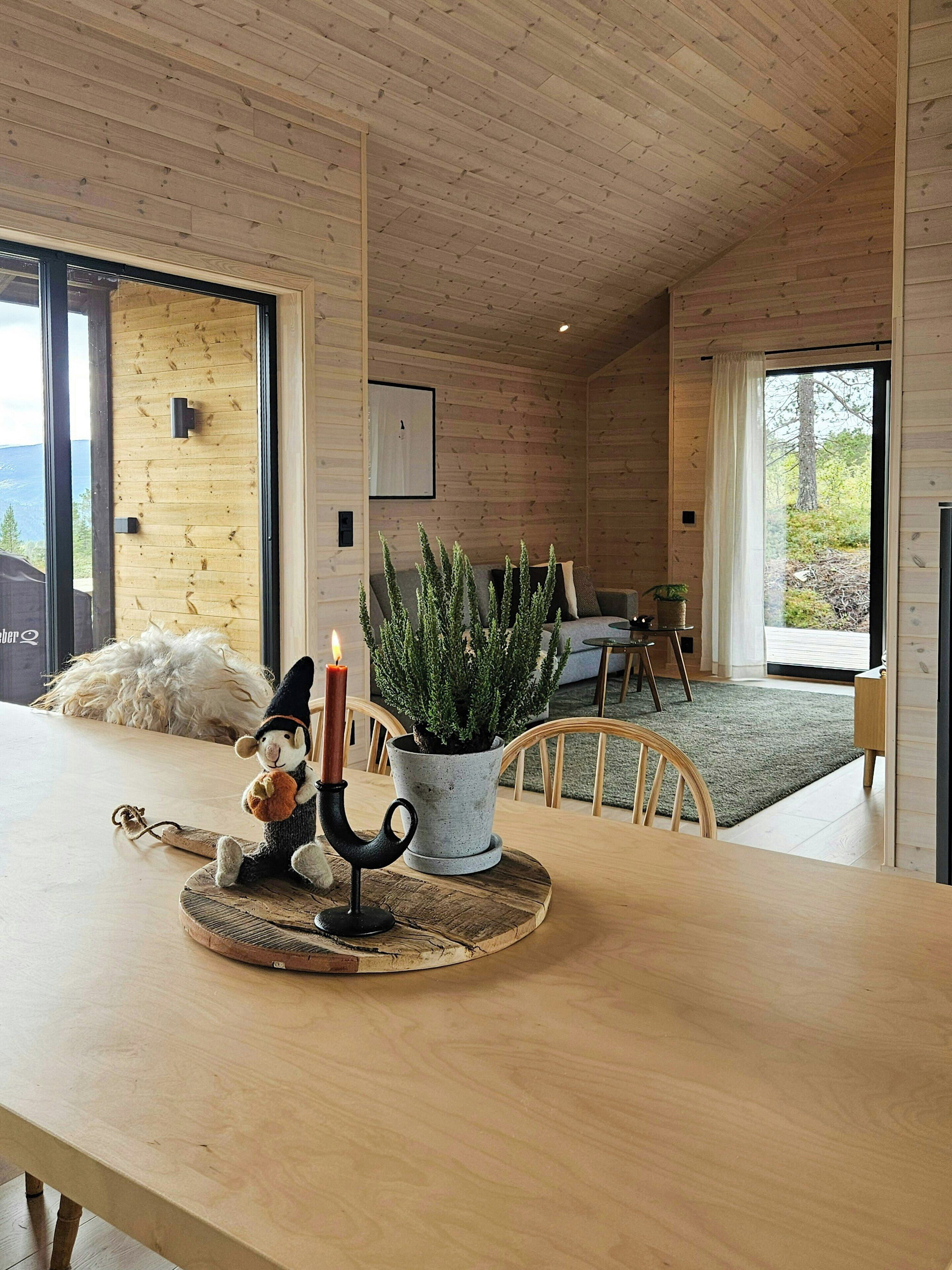 A cozy cabin interior with a wooden ceiling, a dining area with a table, a potted plant, and a decorative candle holder.