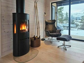 A modern interior with a wood-burning stove, a plush chair, and a large window offering a view of a snowy landscape.