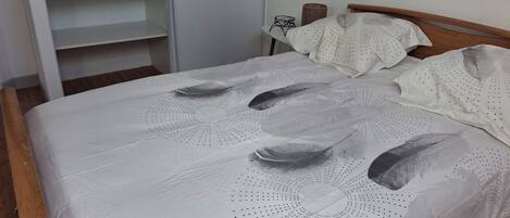 Un lit recouvert d’une couvre-lit blanche ornée d’un motif de plumes, avec une tête de lit en bois et une armoire blanche dotée d’étagères ouvertes.