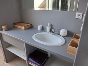 Une salle de bain avec un lavabo blanc, un miroir, un panier en bois, un distributeur de savon et un porte-serviettes.