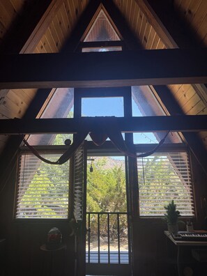 A room with a sloped ceiling, wooden beams, a large window with blinds, and a view of greenery outside.