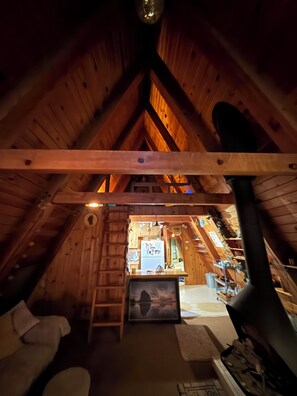 An attic room with wooden beams, a fireplace, a ladder, and a small window.
