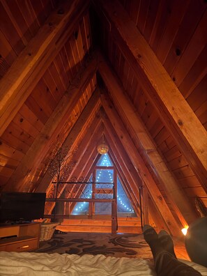 A cozy attic bedroom with wooden beams, a bed, a TV, and a Christmas tree.