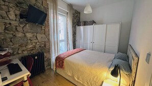A room with a stone wall, a bed with a white headboard, a desk with a computer, a radiator, and a TV mounted on the wall. - Pepe house (Areeta / Las Arenas)