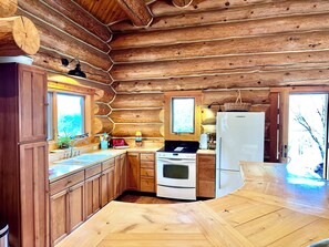 A log cabin kitchen with wooden cabinets, a stove, a refrigerator, and a large window. - Log cabin in the woods with views of Lake MI, Located in historic Cross Village (Harbor Springs)