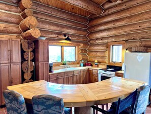 A wooden cabin interior with a wooden dining table, chairs, a kitchen with a refrigerator, and a window. - Log cabin in the woods with views of Lake MI, Located in historic Cross Village (Harbor Springs)