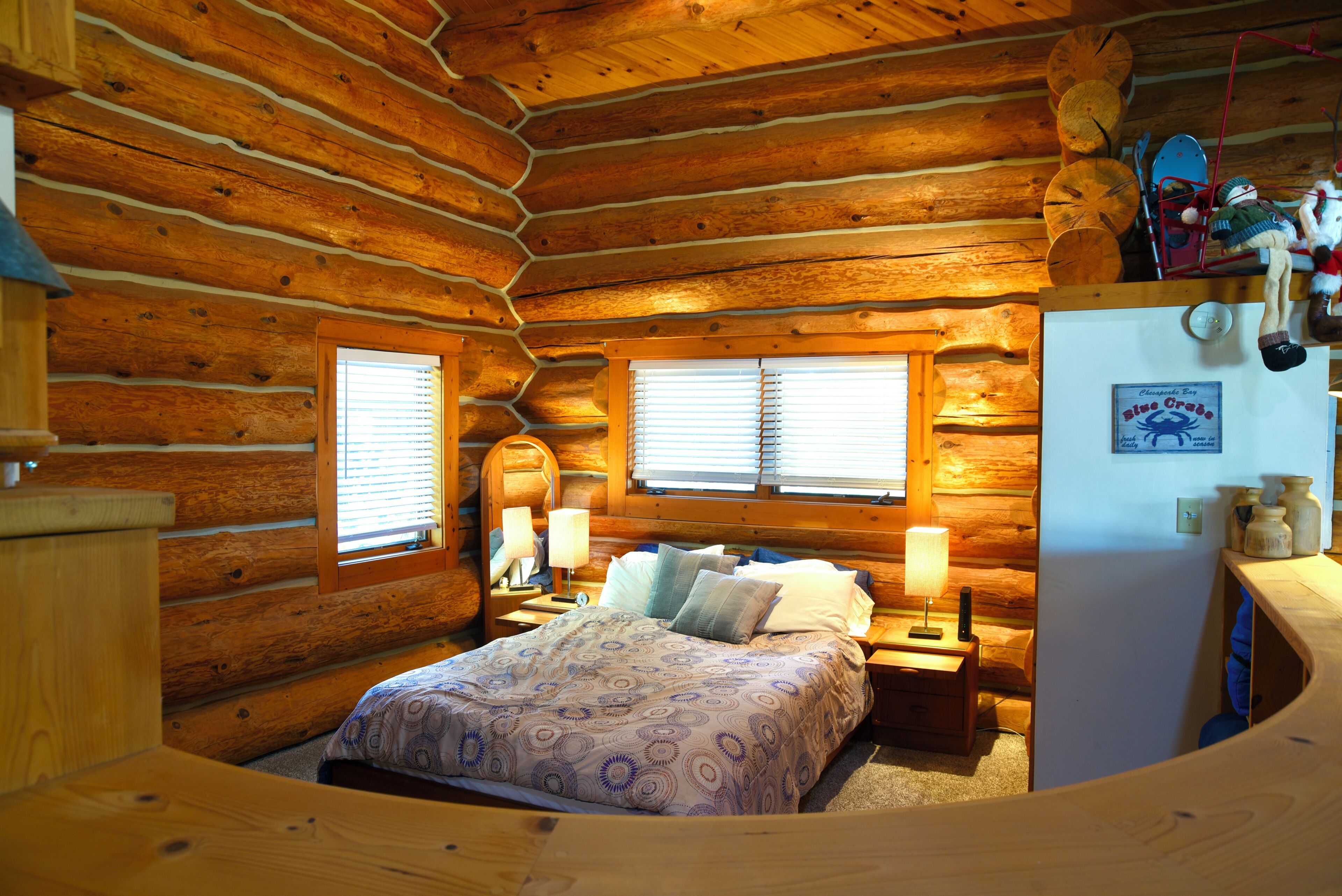 Quarto de cabana de tronco com uma cama, duas mesas de cabeceira, um criado-mudo, uma janela e uma geladeira.