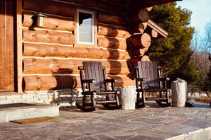 A log cabin with two wooden rocking chairs outside, a stone-paved entrance, and a window with a broken pane. - Log cabin in the woods with views of Lake MI, Located in historic Cross Village (Harbor Springs)