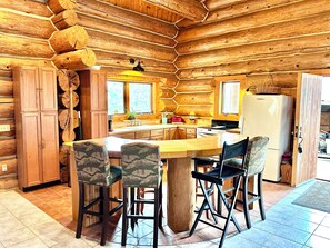 Ein rustikales Blockhaus-Interieur mit einem Holztisch in der Mitte, vier Stühlen, einer Küchenzeile mit Kühlschrank und einem Fenster.