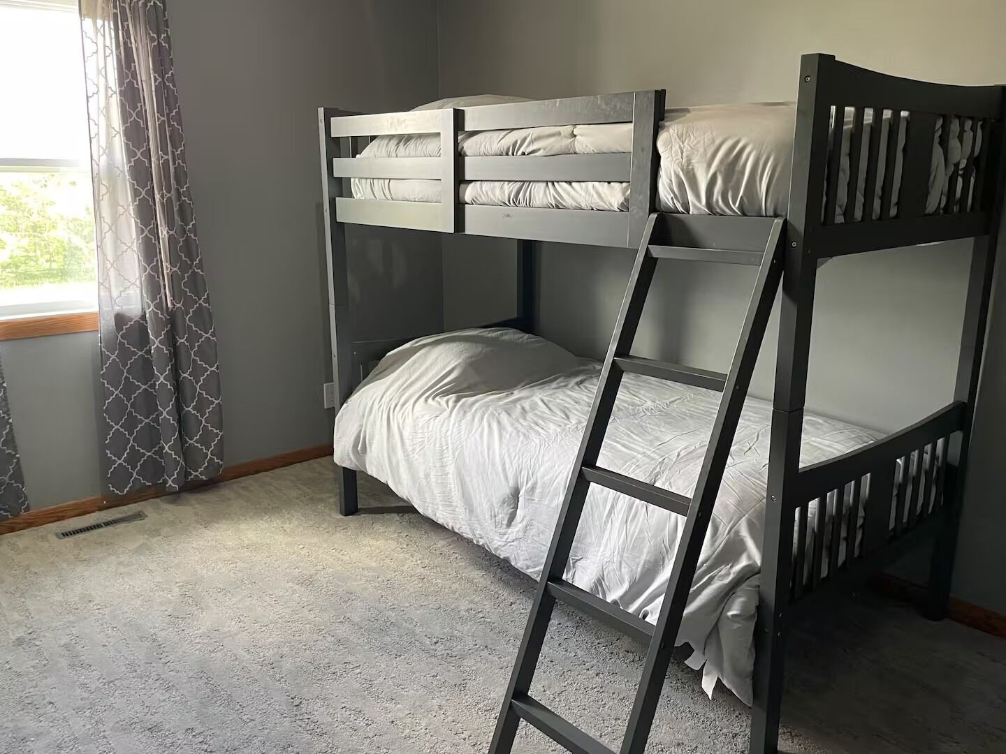 Dormitorio con literas, una escalera, paredes grises y una ventana con cortinas.