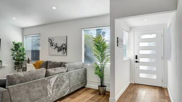 Una sala de estar moderna con un sofá gris, un ventanal con persianas, una planta en maceta y una puerta con panel de vidrio esmerilado.