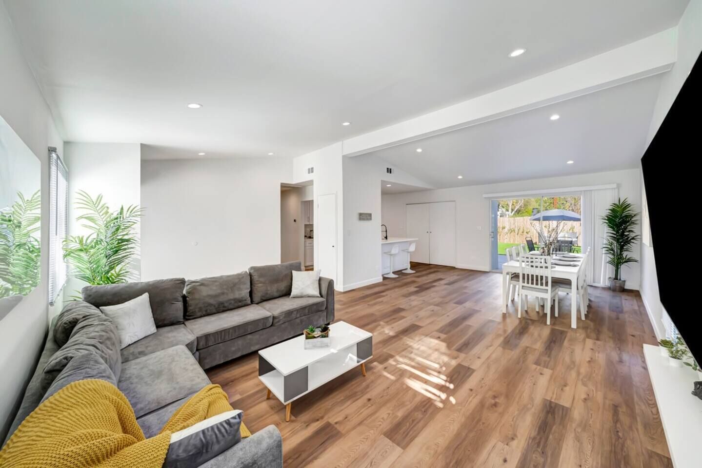 Una sala de estar moderna con un sofá seccional gris, una mesa de centro blanca, piso de madera y un comedor con sillas blancas.