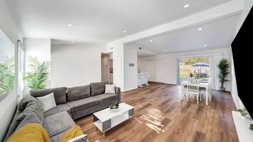 Una sala de estar moderna con un sofá seccional gris, una mesa de centro blanca, piso de madera y un comedor con sillas blancas.