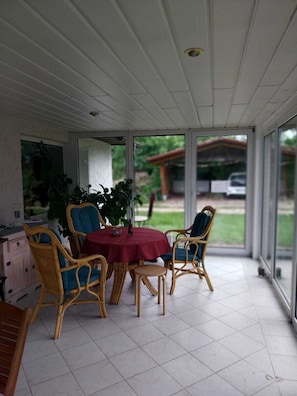 Une terrasse couverte, meublée de meubles en osier, avec une nappe rouge et une vue sur l’extérieur.
