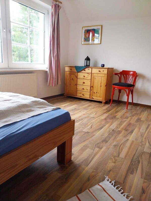 Un dormitorio con cama, cómoda de madera, silla roja y ventana con cortinas.