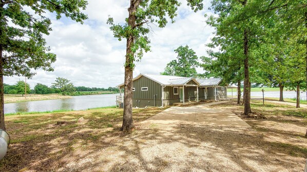 A rustic cabin with a porch, a gravel path leading to it, and a lake in the background.