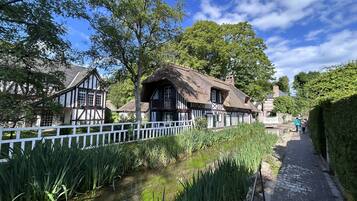 Une maison traditionnelle à colombages, avec un toit de chaume, une clôture blanche et un canal bordé de plantes aquatiques.