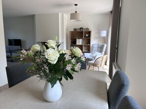 Una mesa de comedor con un jarrón blanco de flores, un sofá gris, una estantería de madera y un sillón gris en una sala de estar moderna.