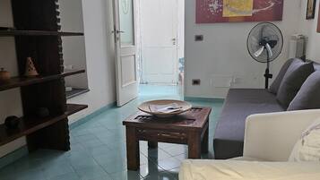 Un salon comprenant un canapé, une table basse, une bibliothèque, un ventilateur et une porte donnant sur une autre pièce.