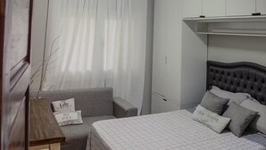 Ein Schlafzimmer mit einem Bett, einem grauen Sofa, einem Nachttisch aus Holz und einem Wandregal.