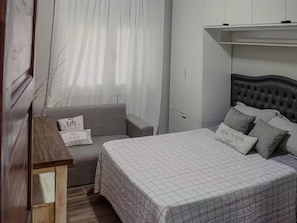 A bedroom with a bed, a grey sofa, a wooden nightstand, and a wall-mounted shelf.
