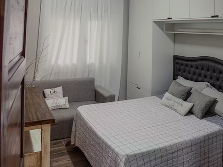 A bedroom with a bed, a grey sofa, a wooden nightstand, and a wall-mounted shelf.. Apartment, excellent location and comfort