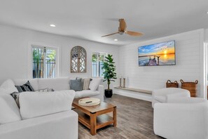 A living room with a white sofa, a coffee table, a potted plant, a ceiling fan, and a sunset painting on the wall. - The Crescent by Duncan Real Estate (Anna Maria)