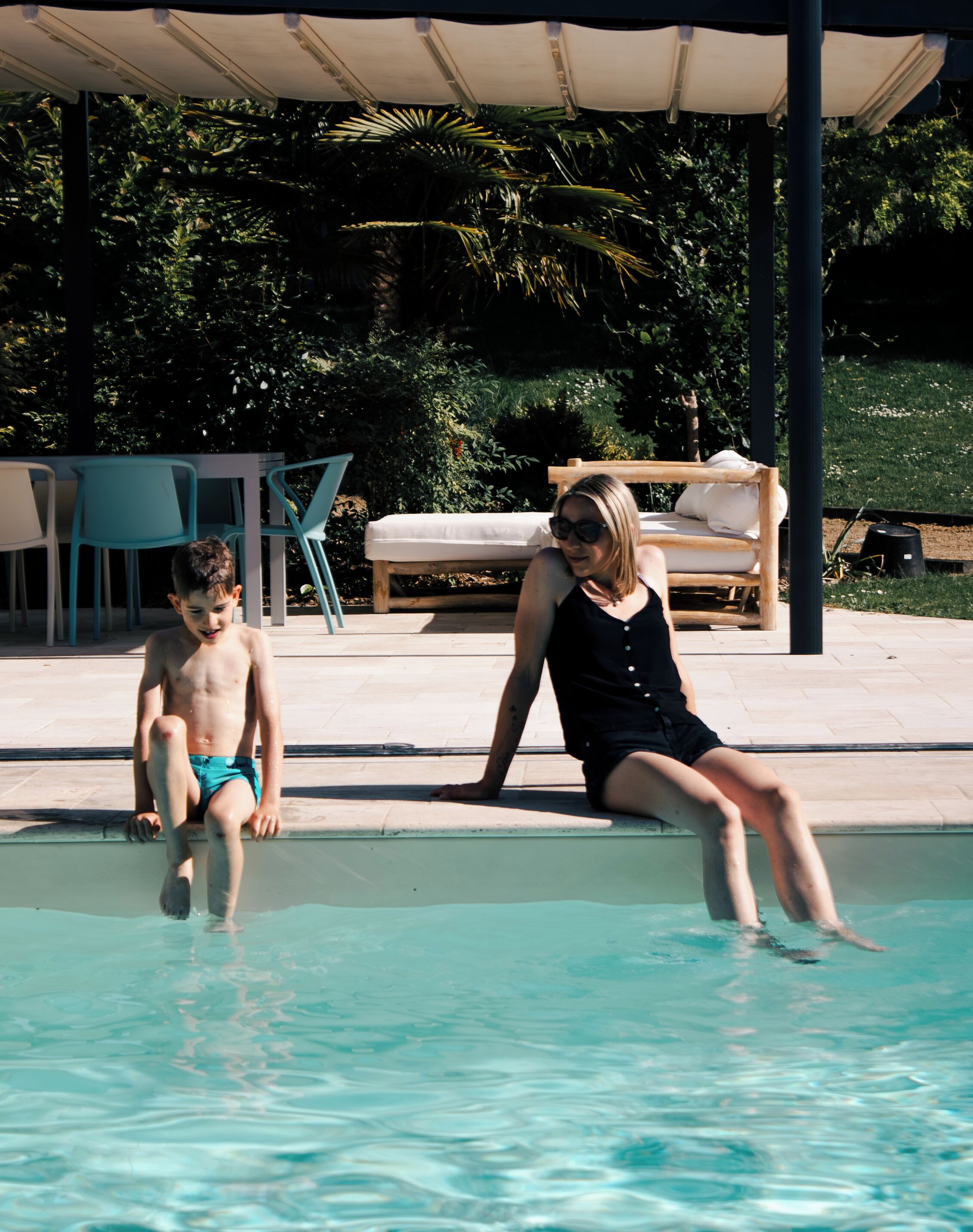 Un área de piscina con un banco y sillas de madera, una zona de estar techada y una piscina con agua cristalina.