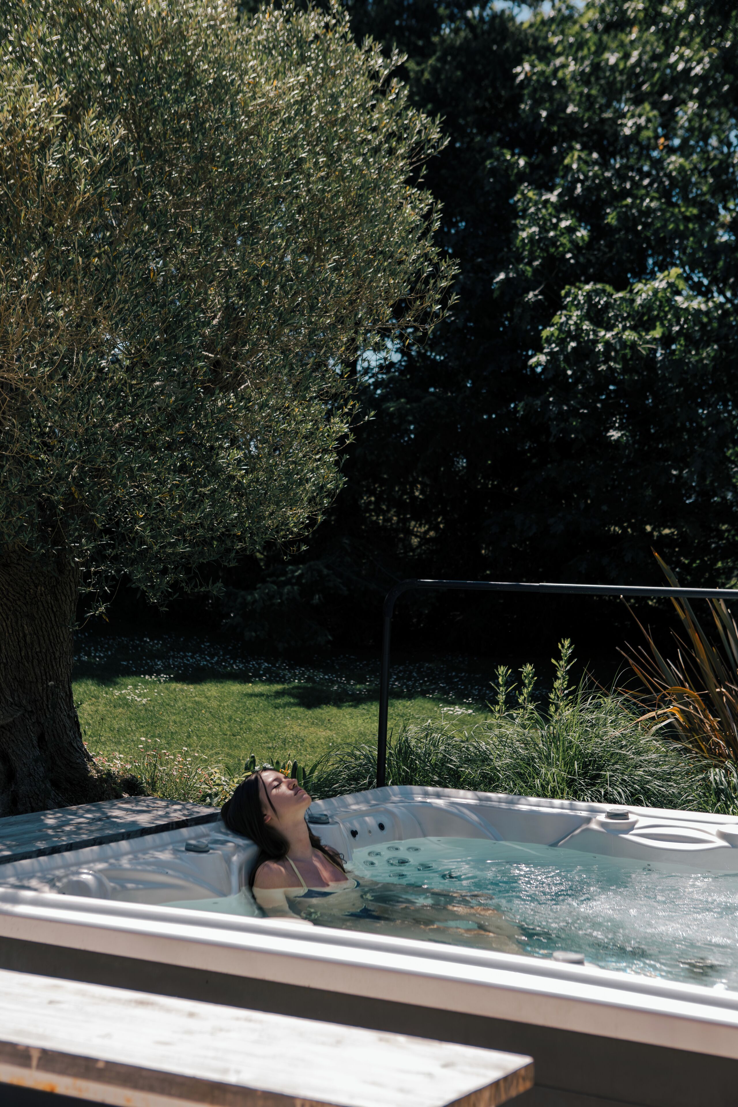 Een jacuzzi met daarin iemand ontspannend, omringd door groen en een boom.