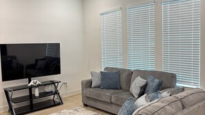 Ein Wohnzimmer mit Flachbildfernseher, zwei grauen Sofas, Deckenventilator und großen Fenstern mit Jalousien.