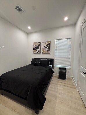 A bedroom with a bed, a bedside table, a window with blinds, and two wall-mounted artworks.