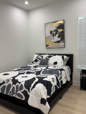 A bedroom with a bed, a wall-mounted abstract painting, and a window with blinds.