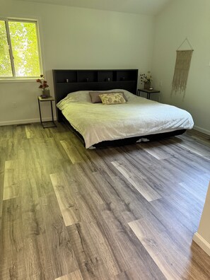 Un dormitorio con cama, suelo de madera, una ventana y un macramé colgado en la pared.