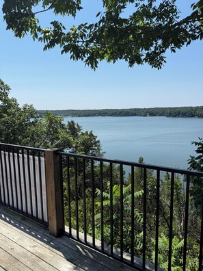 A wooden deck with a metal railing overlooking a calm lake with dense foliage and a clear sky. - Deer Run on Eddy Bay
4 bd , 3 bath luxury lake living! (Eddyville)