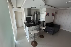 Una cocina compacta con electrodomésticos modernos, una zona de comedor con un taburete de bar y un sofá gris.